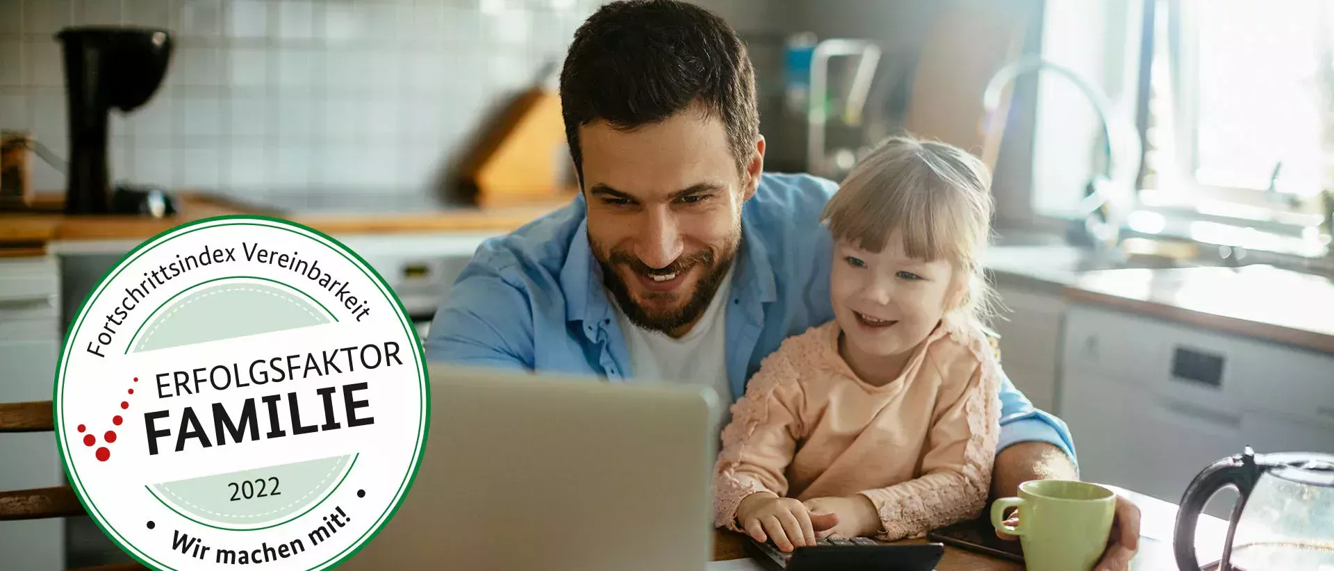 Father sits at the kitchen table with his young daughter and they look happily at the laptop in front of them.